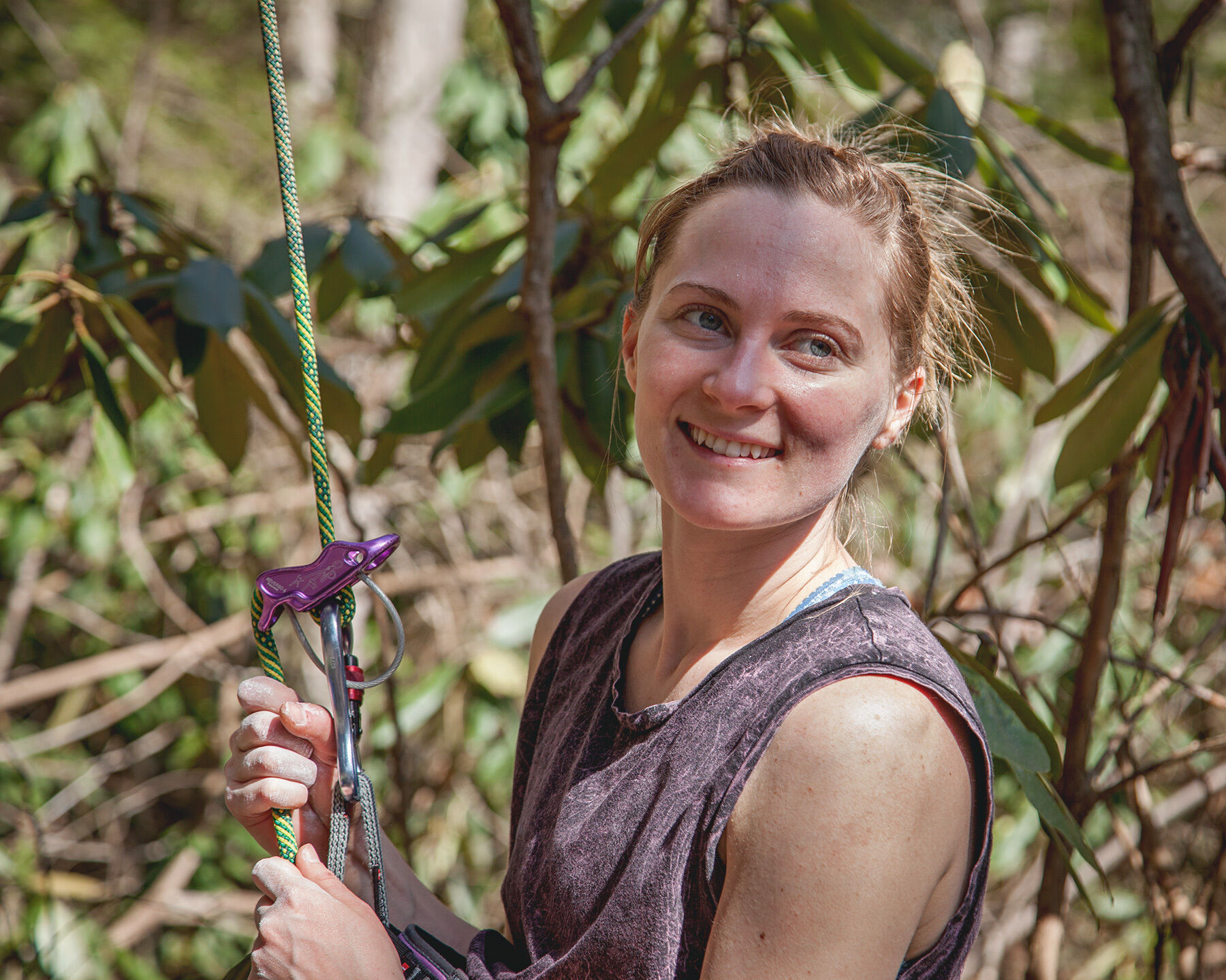 Landscape-oriented color photograph. A tight semi-profile shot of a woman holding onto a green climbing rope in a purple belay device smiles and looks off frame to the right. She has blond hair pulled up in a ponytail, fair skin, and blue eyes, and is wearing a gray tank top. The image is cropped at her torso, and her hands are covered in chalk. The sun is bright and casting half of her face and torso in shadow. All around her in the background are the leaves and branches of trees and bushes, slightly out of focus.