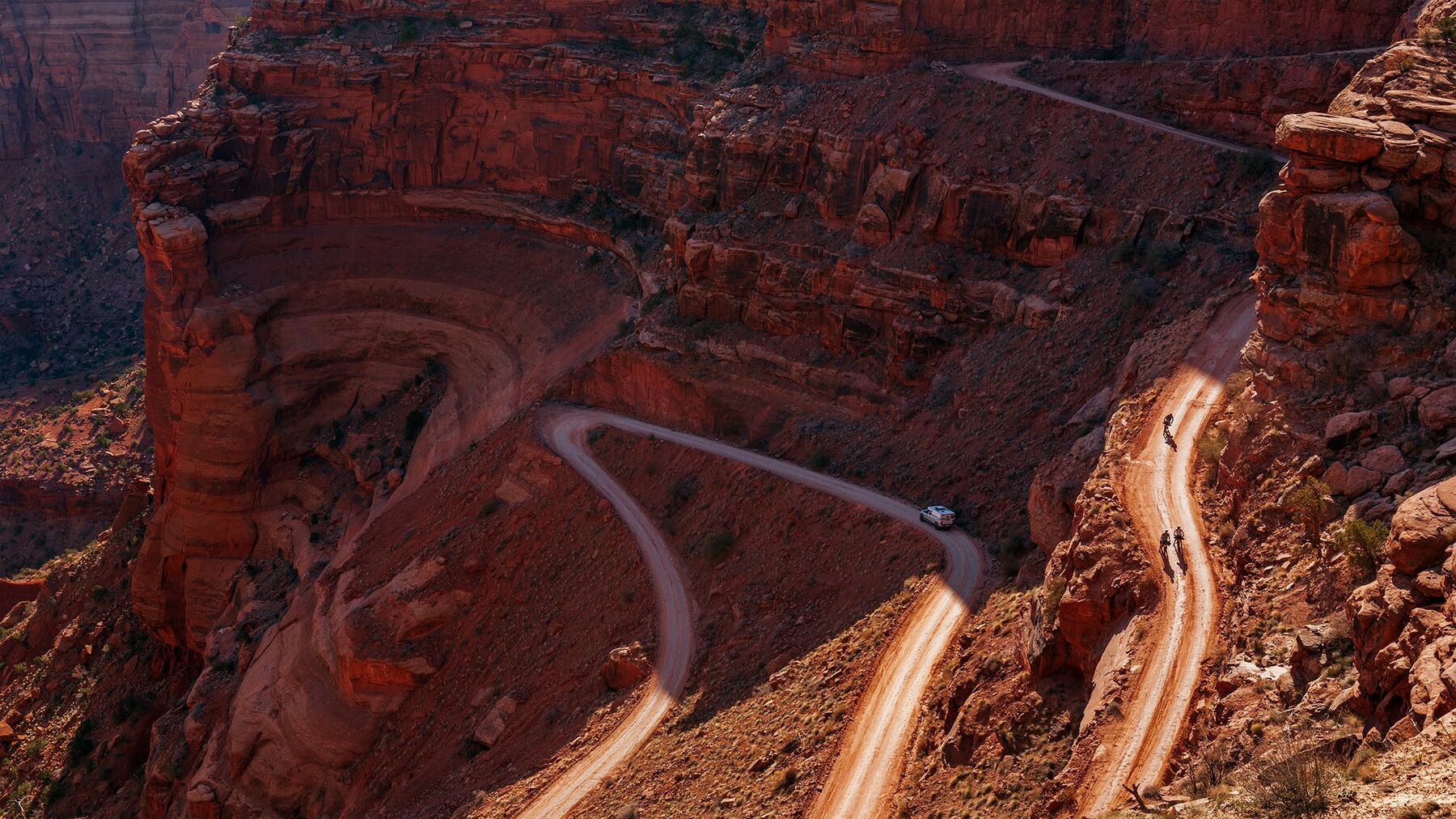 A landscape-oriented color photograph taken from a high vantage point showing a winding dirt road descending a red sandstone canyon in the desert. The road starts in the top right and zig-zags toward the bottom left. Three cyclists can be seen near the top of the road, and a truck is half-way down. The warm desert sun is illuminating the lower-right diagonal.