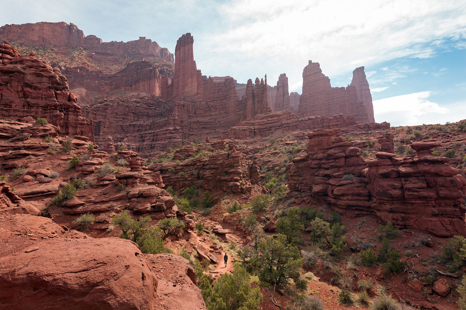 Landscape-oriented color photograph. A sprawling expanse of sandstone towers and rock formations stretches from just below the top of the frame at the left towards the middle of the frame at the right. Started at the bottom left of the frame a hiking trail can be seen in the distance, leading towards the center of the frame, where a lone hiker can just be made out on the trail. The sandstone is varying shades of brown and red, and the landscape is sprinkled with green sage and juniper bushes. At the center of the frame near the top half the tallest, proudest desert towers stand like skyscrapers against the light blue sky, which is filled with soft white clouds. The sun is somewhere above the frame, creating a slight haze towards the top of the frame.