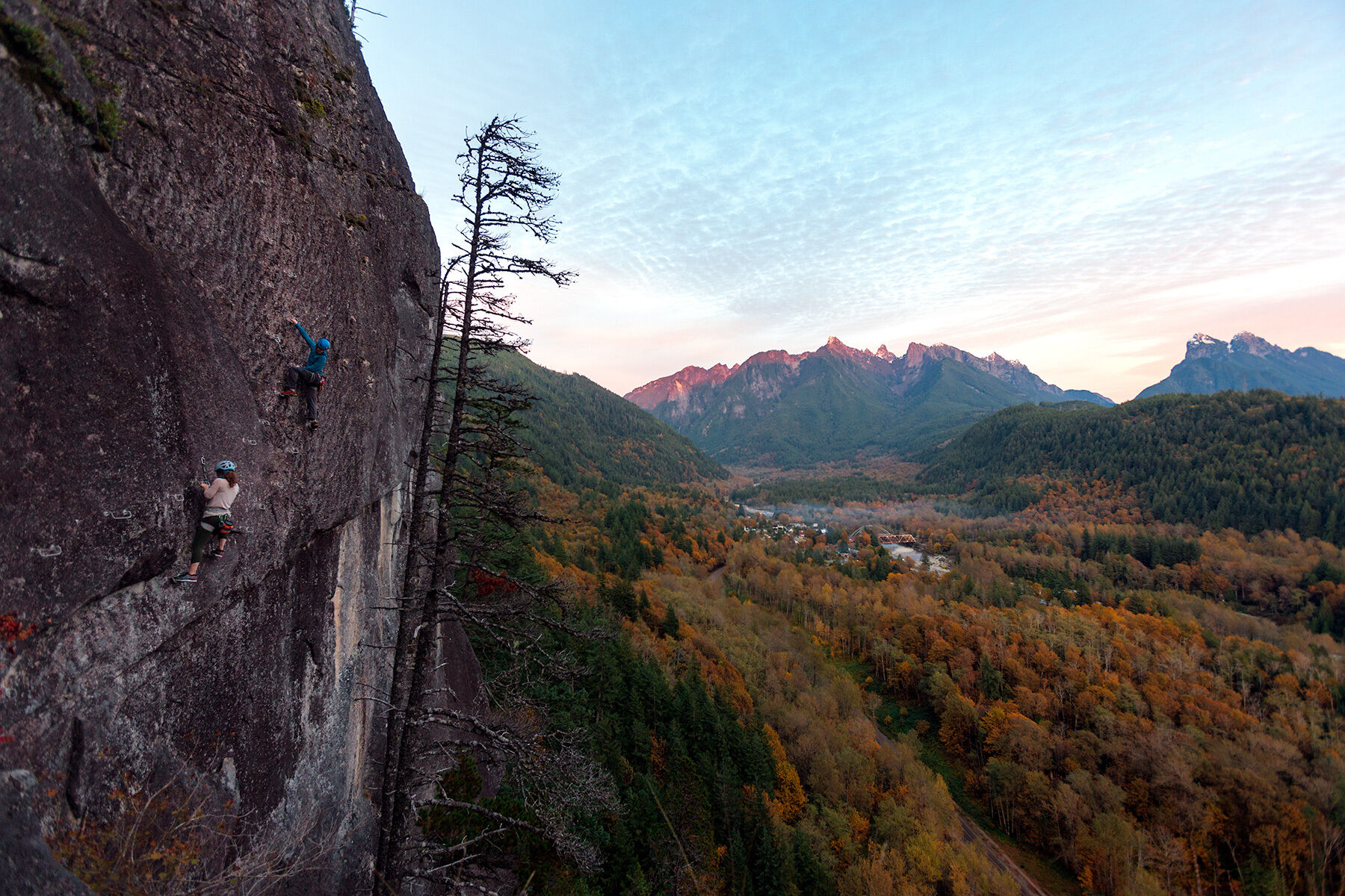 Lanscape-oriented color photograph. A wide shot taken from high up on the side of a rock cliff overlooks a vast landscape of trees leading to a mountain range in the distance. The left third of the image is filled with a vertical wall of dark rock, with streaks of lighter gray colored granite throughout. Just visible are iron ladder rungs on the side of the rock face leading from the bottom left of the frame towards the top of the rock cliff. Two rock climbers are seen in action ascending the rock face. The climber closer to the left edge of the frame is in a light-colored long-sleeve shirt, with long curly hair protruding from her light-gray helmet. The second climber is a little farther and higher than the first and is in a long bright blue jacket and bright blue helmet, and is hanging looking down towards the first climber. A tall, mostly bare tree separates the rock wall from the rest of the landscape. The sea of trees is a smattering of dark and light green with bright yellow and orange-colored trees sprinkled among them. Near the center of the expanse some buildings and smoke or fog can just be made out. The tips of the mountain range have a small amount of snow and are glowing pink from the light of the setting sun. The sky is a bright blue with splashes of pink and orange near the horizon.