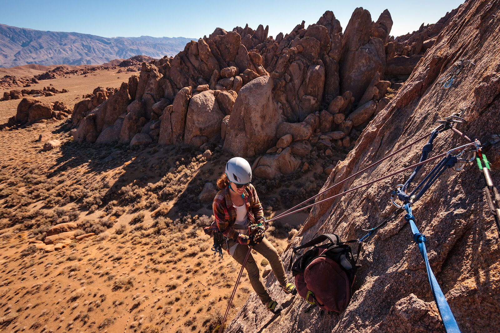 Landscape-oriented color photograph. A wide shot taken from the side of a jagged face of a rock wall looking down on a woman in a light-gray helmet, sunglasses, and plaid shirt, smiling as she rappels down the rock face. The woman is horizontally centered in the frame, and is rappelling towards the bottom of the frame, and she is facing towards the right, where the rock face is. Below her is a desert floor filled with sand, small desert shrubs, and boulders. Seen above and behind her are large clusters of giant jagged boulders and rock formations that build toward the upper right of the frame. In the distance a pale blue sky can be seen just below the top edge of the frame and towards the left in the distance is a rolling mountain range. The sunlight in the image is harsh and the giant rock formations behind the woman are casting deep harsh shadows on the ground below. In the bottom right corner of the frame there are a couple of different types of rope riggings tethering the photographer, who is out of frame, to an anchor from which the woman is rappelling. A small backpack is affixed to this anchor and is hanging on the side of the rock face as well..