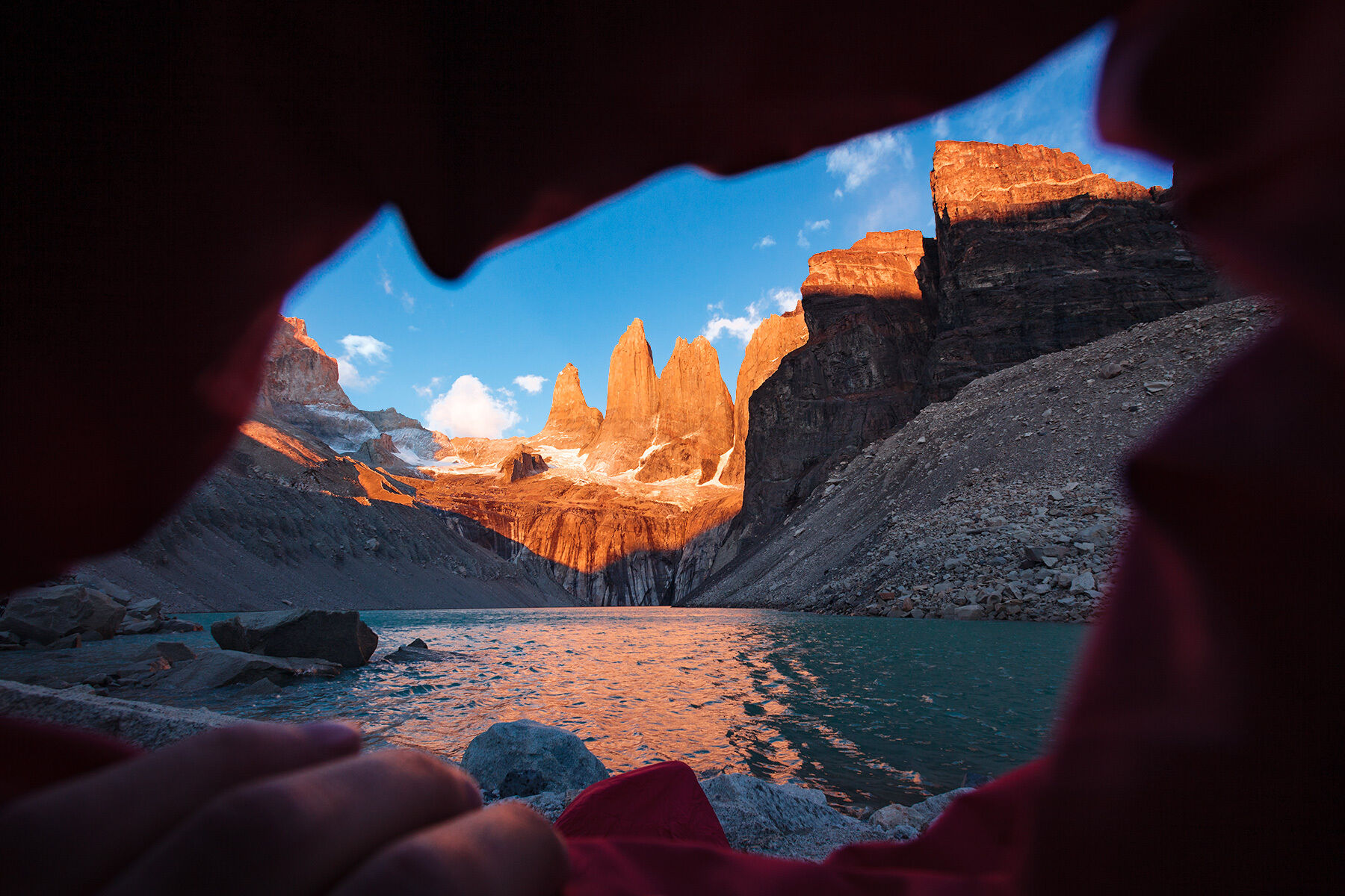Landscape-oriented color photograph. A very wide shot from the perspective of inside a sleeping bag, the edges of the opening of which frame the entire shot, a deep red, with a left hand holding open the bottom edge. The feet of the person in the sleeping bad can be seen just in front of the opening at the bottom center of the frame. The scene beyond is a broad landscape of a set of three tall, prominent rock towers piercing a brilliant blue sky at the head of a small body of water which stretches out toward the feet of the sleeping back. There is a broad streak of bright orange sunlight coming from out of frame at the left illuminating the towers and the tips of some surrounding rock, and the middle ground is in cool shadow. A band of bright orange and pink streak across the center of the body, reflecting off the towers at the opposite end. Some small patches of snow can be seen at the base of the towers, and a handful of soft white clouds are in the sky beyond.