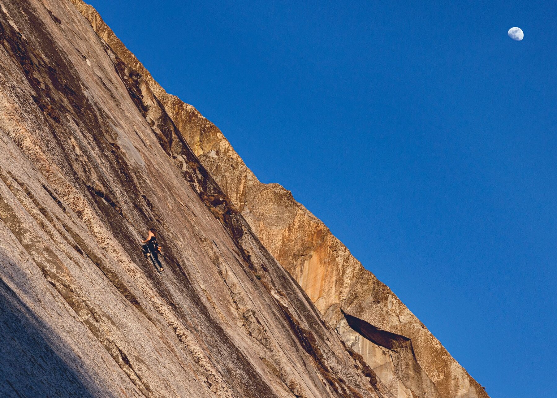 Landscape-oriented color photograph. A massive granite slab of rock fills the left side of the frame at an angle from the top left to the bottom right, which has streaks and scars of light gray, dark gray, and shades of orange. The right side of the frame is filled with a clear, deep blue sky, and a small three-quarters moon shines bright in the top right corner. The bottom left corner has a streak of deep shadow covering it. A female rock climber can be seen half-way up the rock face at a distance, mid-climb.