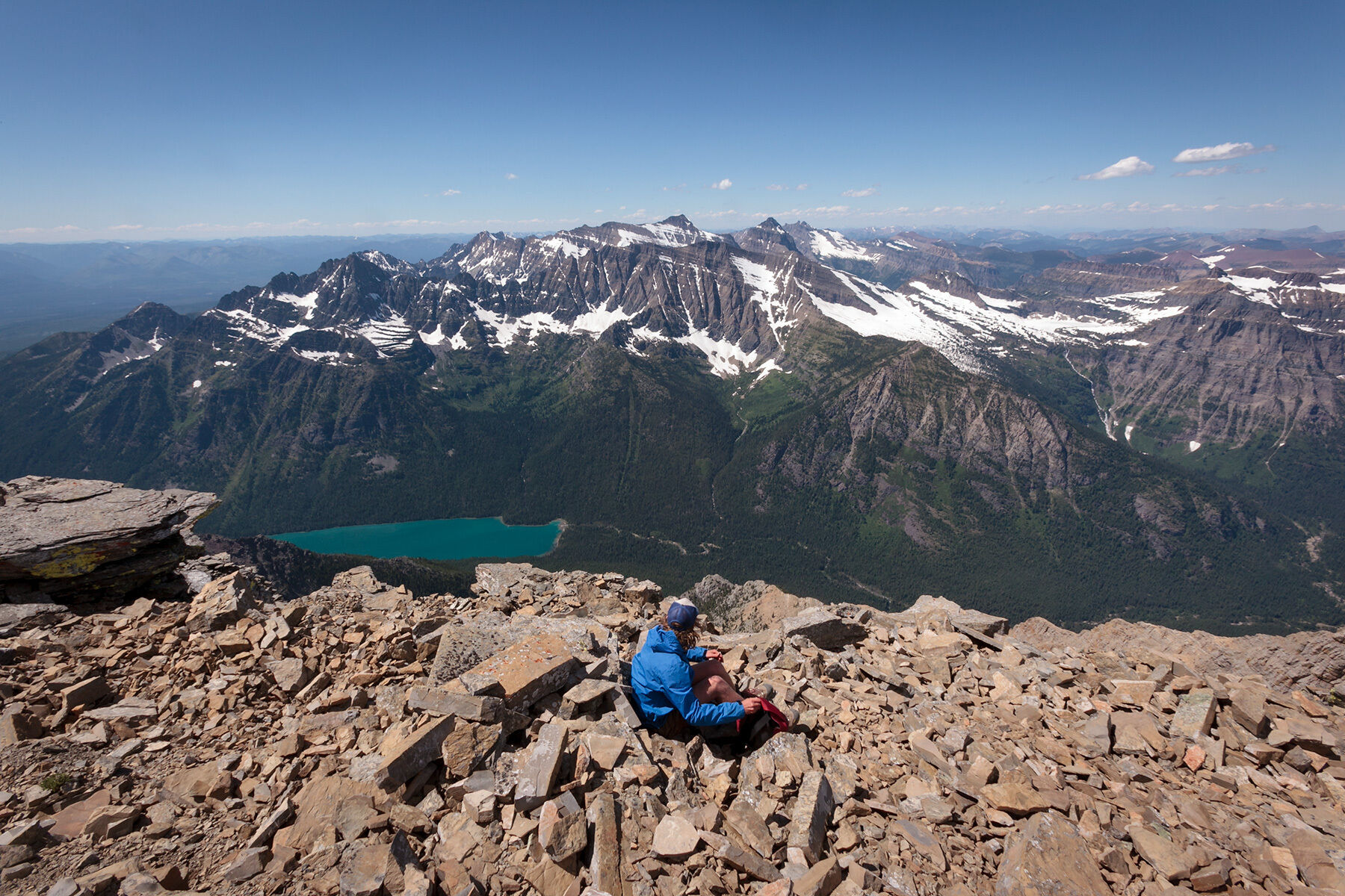 Douglas sitsatop Carter Peak in Glacier National Park, Montana with Bowman lake and snowcapped mountains in the background