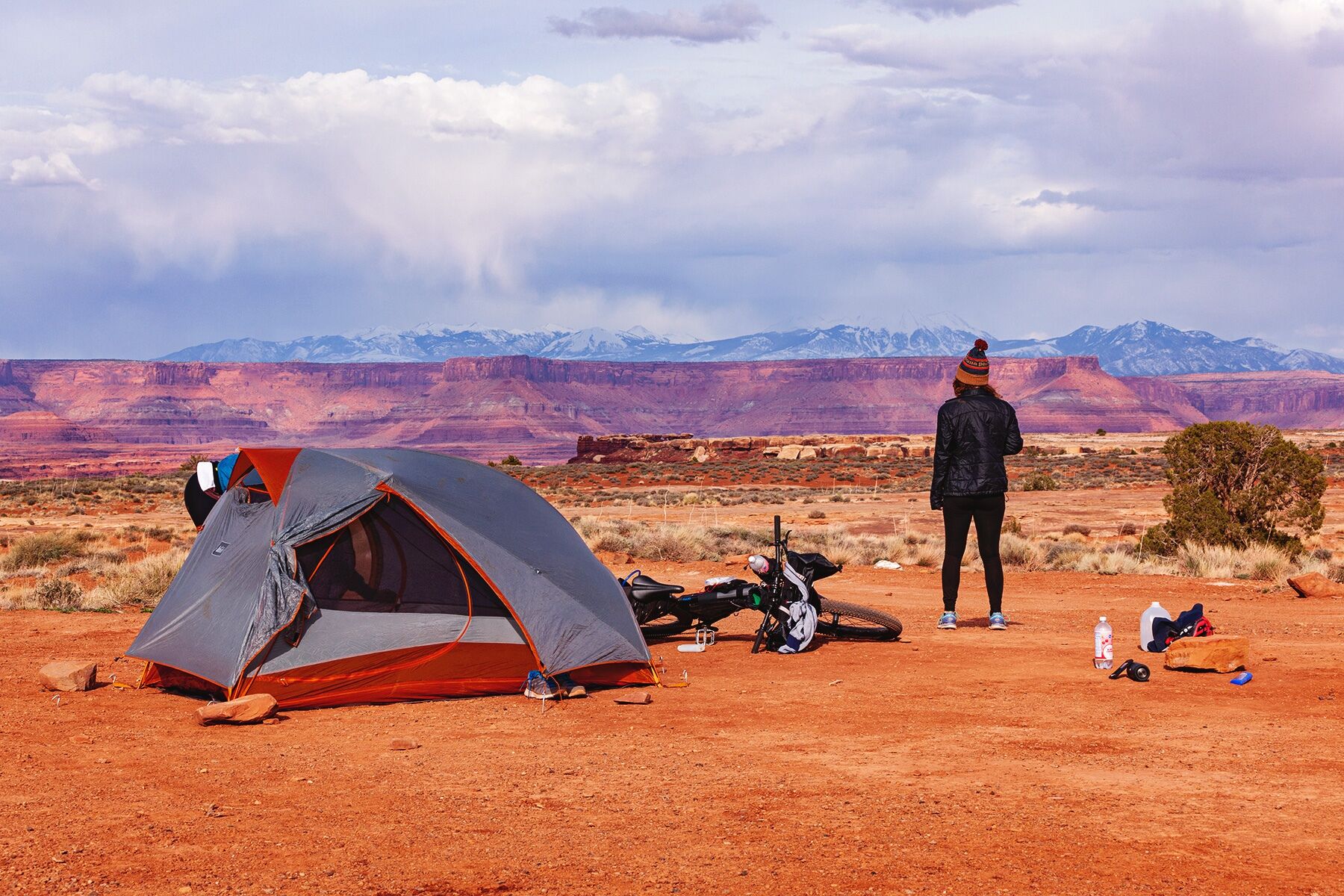 A landscape-oriented wide shot showing a campsite in the foreground, and sandstone cliffs, snow-capped mountains, and tick clouds in layers in the distant background. There is a tent on the left side of the foreground, a mountain bike laying on the ground to the right of it with various camping items strewn across the ground, and then a woman dressed in black leggings, a black jacket, and a navy and orange winter hat, her back to the camera, looking off into the distance.