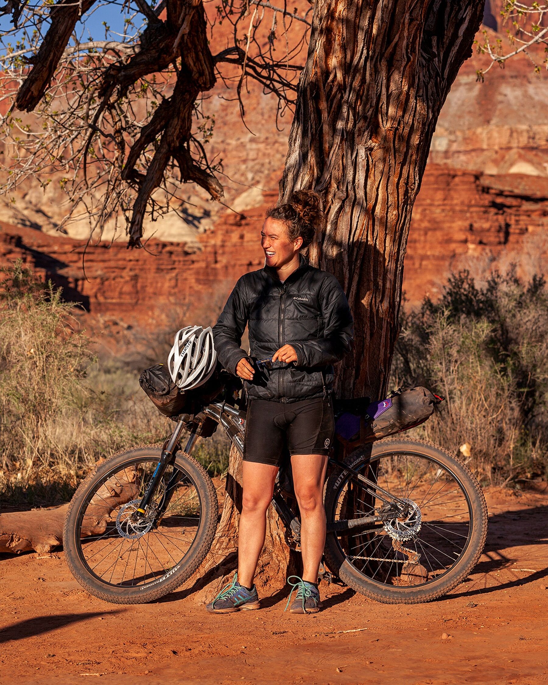 A portrait-oriented photograph showing A woman in cycling attire standing in front of a bike loaded up with bikepacking gear looking off-frame to the left, smiling. The bike is leaning up against a large tree and bushes and sandstone rock formations can be seen in the background. The red earth in the foreground, the woman, and the surroundings are glowing in the warm morning sun.