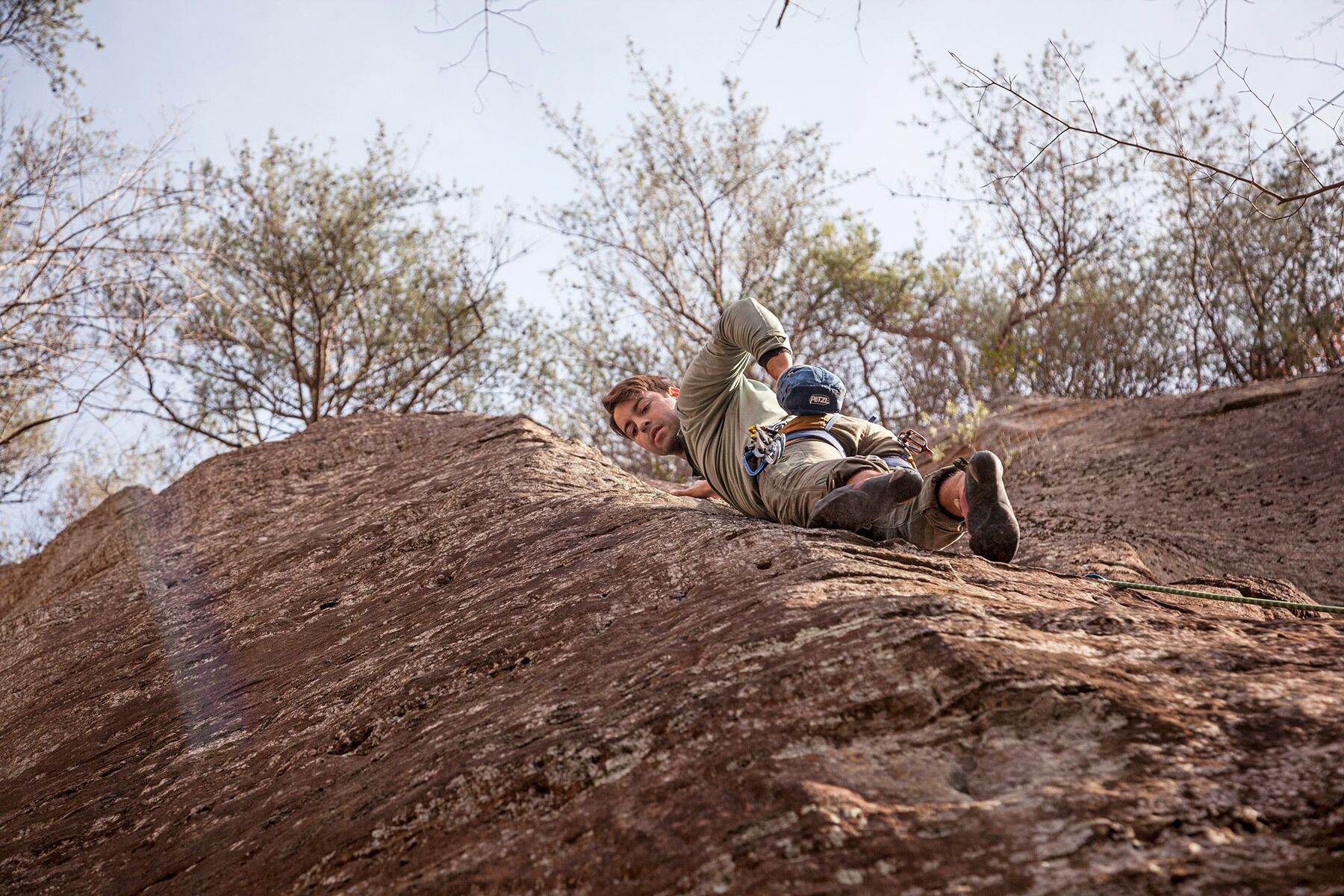 Landscape-oriented color photograph. A low shot looking up at a rock climber who is balanced on the side of a rock face by only his toes and his right hand, which is gripping the rock above him. He is looking down and to the left at his next foot placement and his left hand is placed in his chalk bag, which is attached to his harness at his waist. He is positioned on the right side of where the rock face meets a soft edge that slopes at a different angle toward the left, and the left side of the rock face is in slight shadow. The man is positioned in the center of the frame, and above him the rock ends and meets a row of trees hanging over the cliff's edge, and beyond them a light blue sky is visible. The afternoon sunlight is soft but streams across the right face where the man is climbing, highlighting a climbing rope that can just be seen coming from below the climber and out of frame to the bottom right.