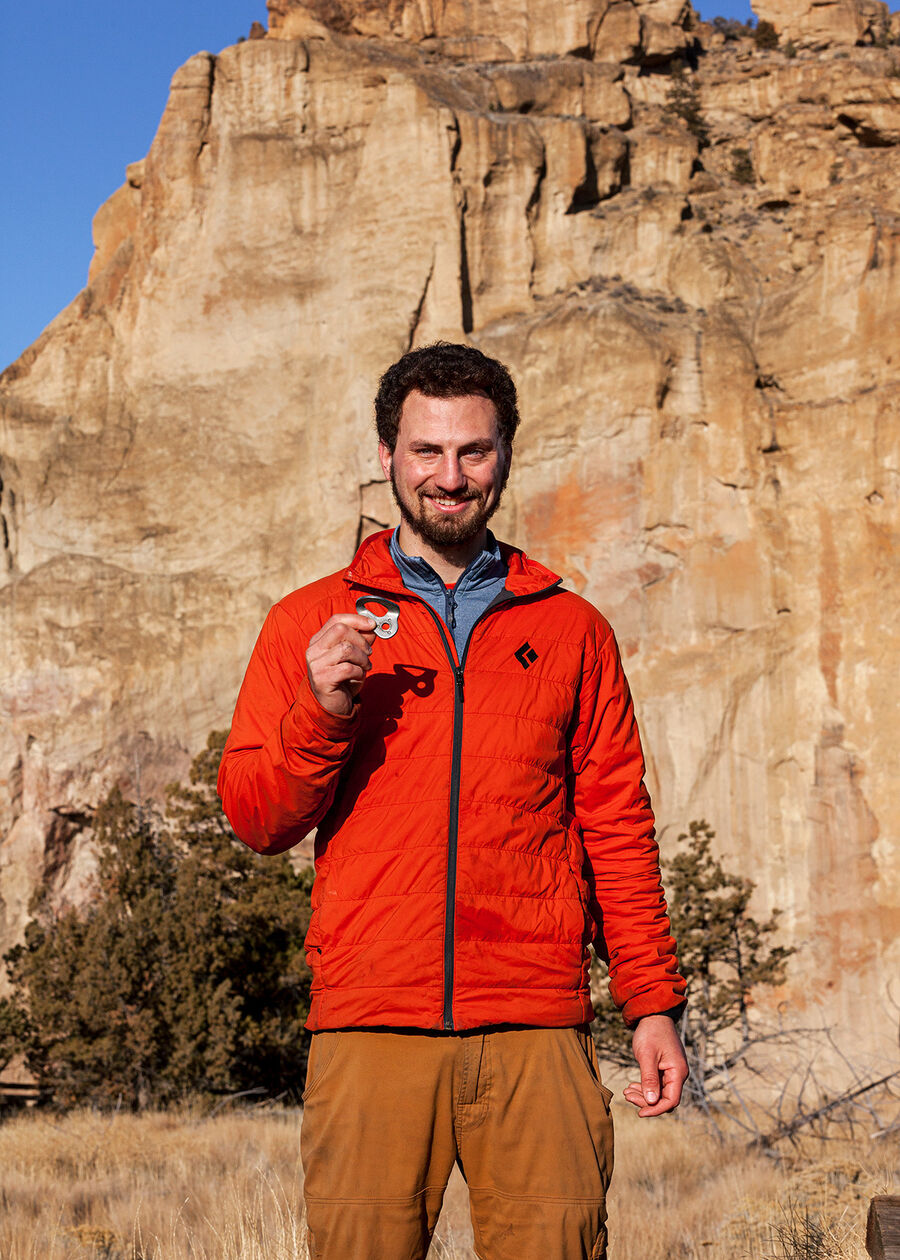 Portrait-oriented color photograph. A man in a bright orange jacket and orange-brown pants stands in the center of the frame, cropped at the knees, his head just above the center of the frame vertically. He is in sharp focus, and behind him the distance is a massive orange and brown rock face that almost fills the entire frame except for some small areas revealing a bright blue sky, slightly out of focus, and some brown grass and a green bush slightly less out-of-focus in the middle ground. He has fair skin, medium-length brown, tight curly hair, and a short beard. He's smiling and looking directly at the camera and holding a small piece of metal up in his right hand, which is casting a dark shadow on his torso from the direct harsh sunlight.