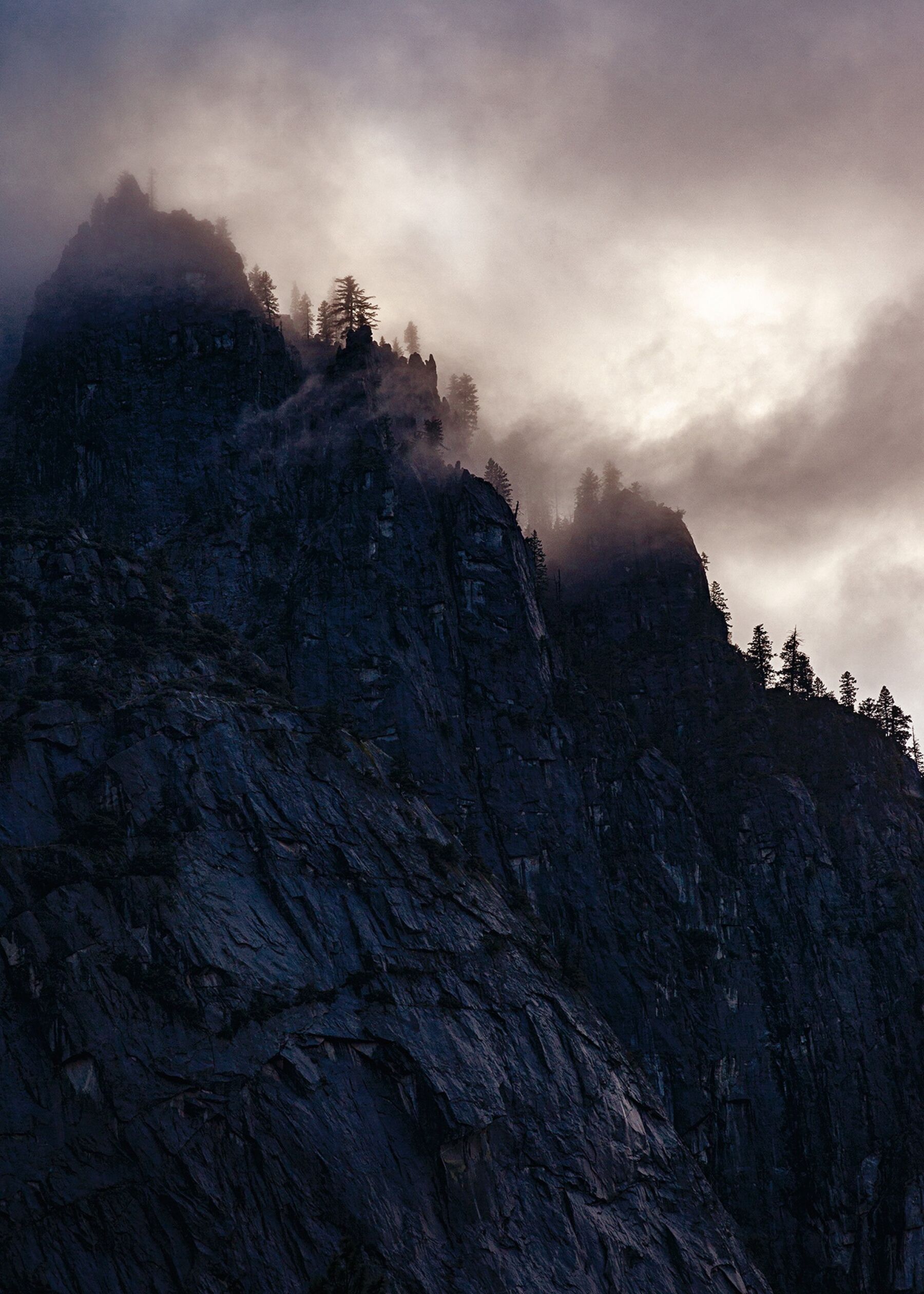 A dark, cloudy sky hovers over the silhouette of a tree-topped cliff in Yosemite National Park, California