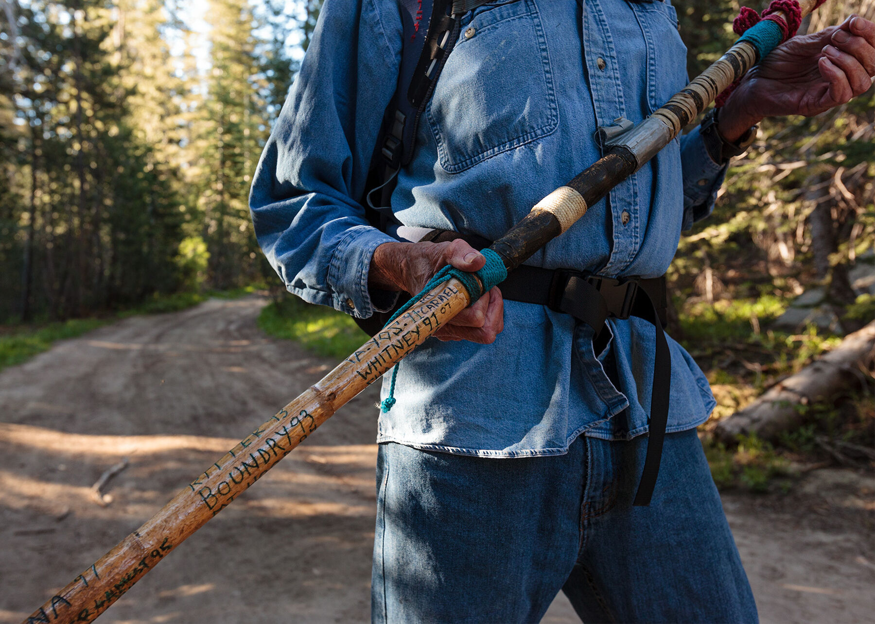Bill standing on a trail holding up his hiking stick with carvings of peaks he has summited carved on the side