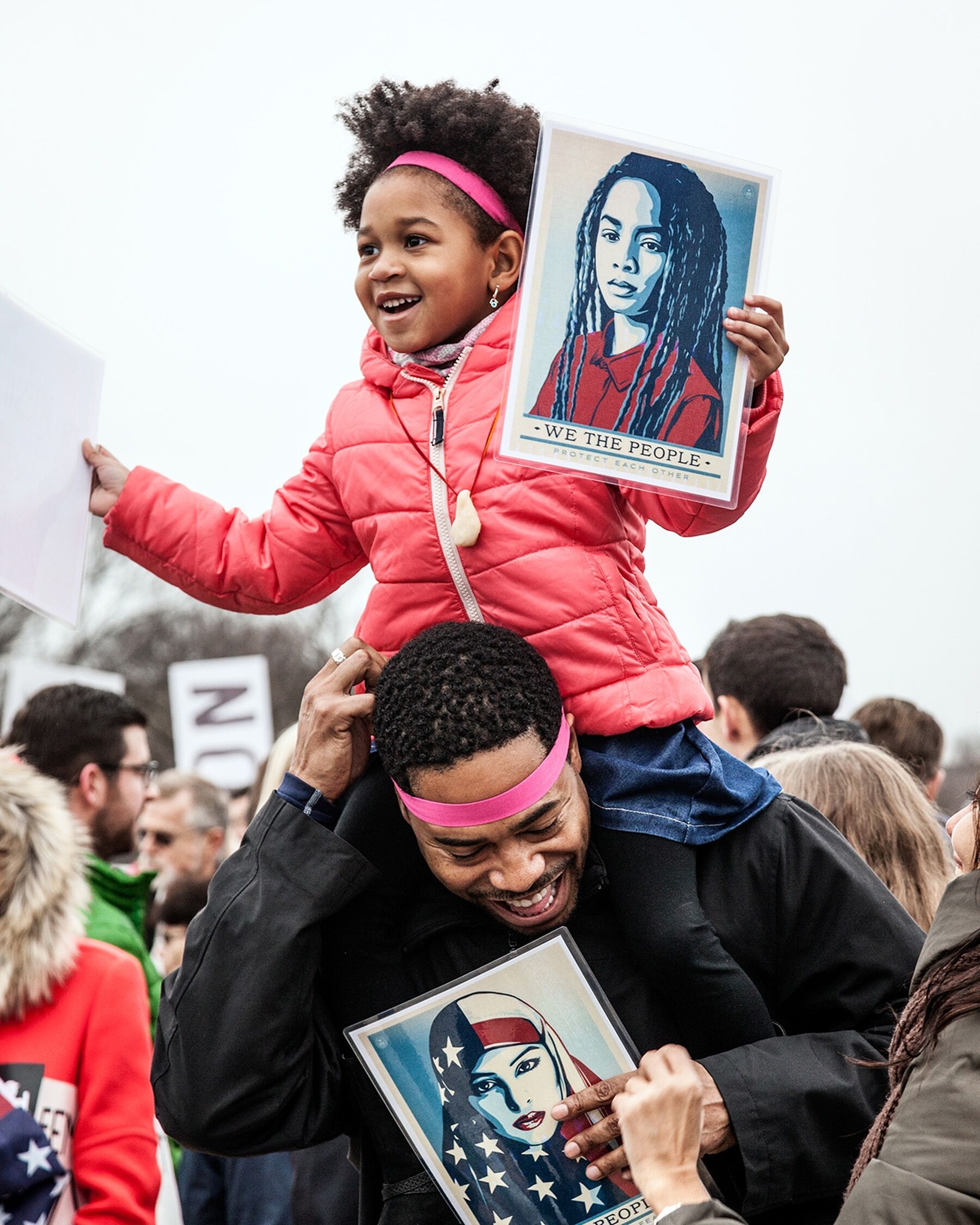 A young girl in a pink jacket sits on the shoulders of a man at the Womens March on Washington in 2017