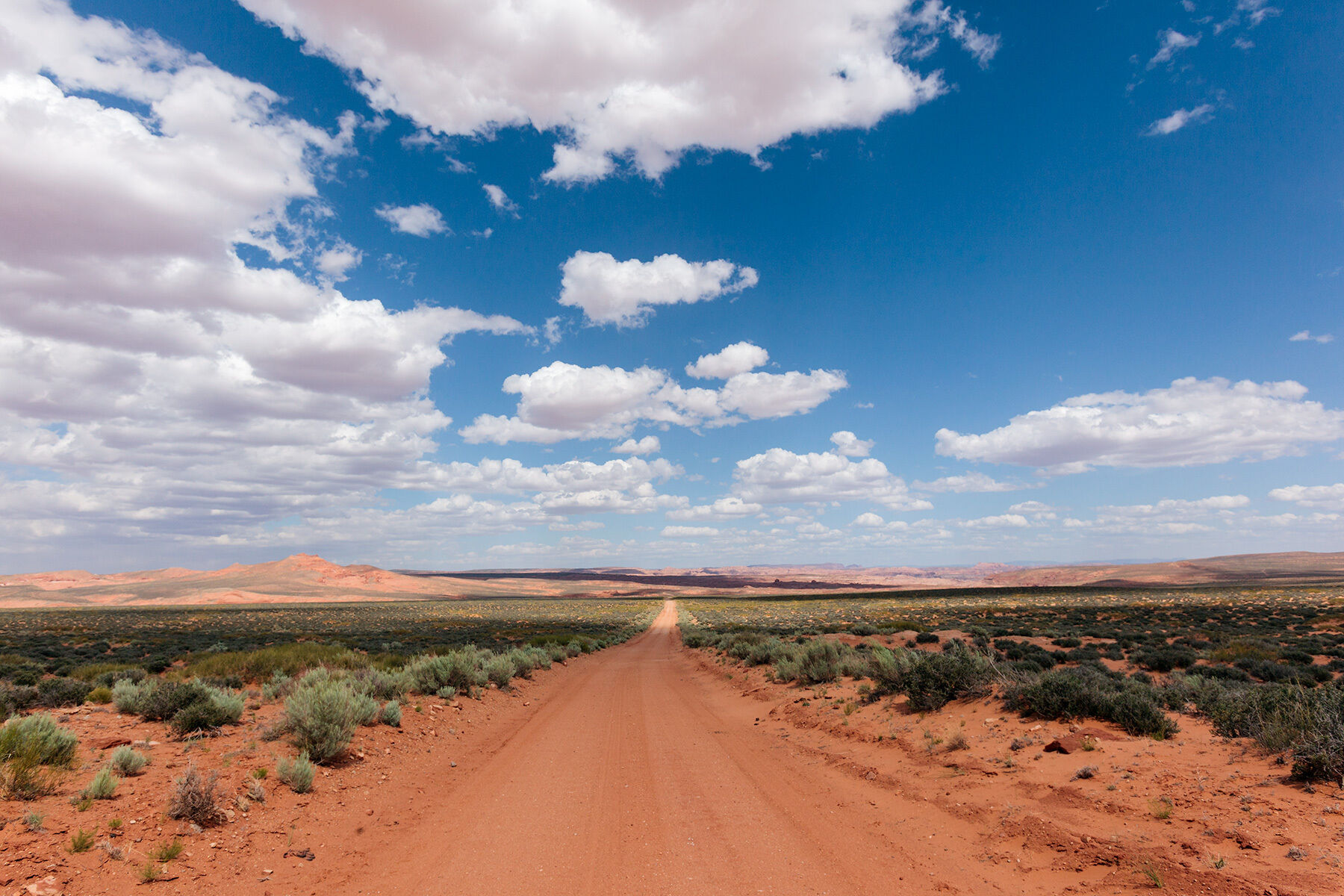 A deep blue sky with big fluffy white clouds, sandstone mountains in the distance, looking down a long desert dirt road in Southern Utah